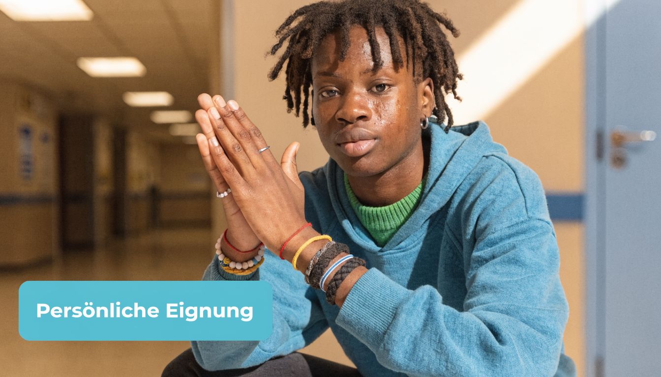 Ein junger Mann mit Dreadlocks und blauem Kapuzenpullover sitzt nachdenklich in einem Flur vor dem Text Persönliche Eignung.
