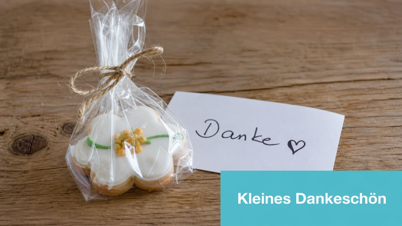 Ein in Zellophan verpackter Blumenkeks neben einer kleinen Dankeskarte mit handschriftlichem Dankeschön auf einem Holztisch.