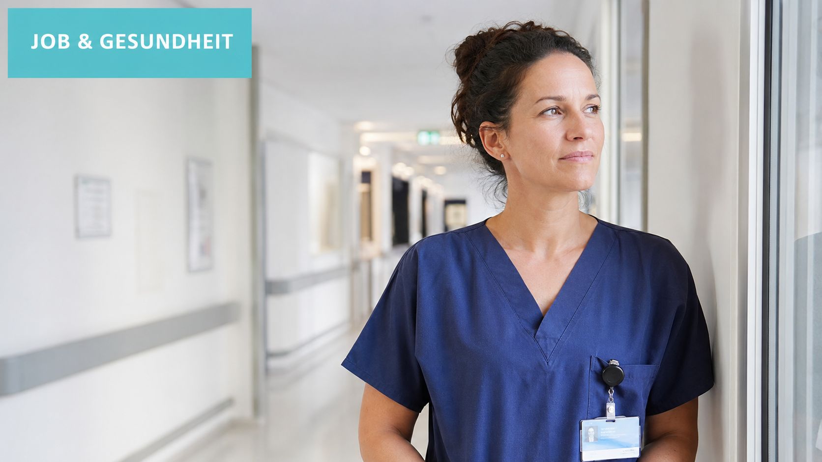 Eine nachdenkliche Krankenschwester in dunkelblauer Arbeitskleidung steht in einem hellen Flur eines Krankenhauses und schaut aus dem Fenster.